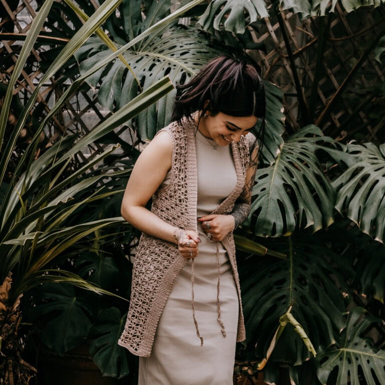 A young women wearing a long crochet vest over a knit dress standing in front of large greenery.