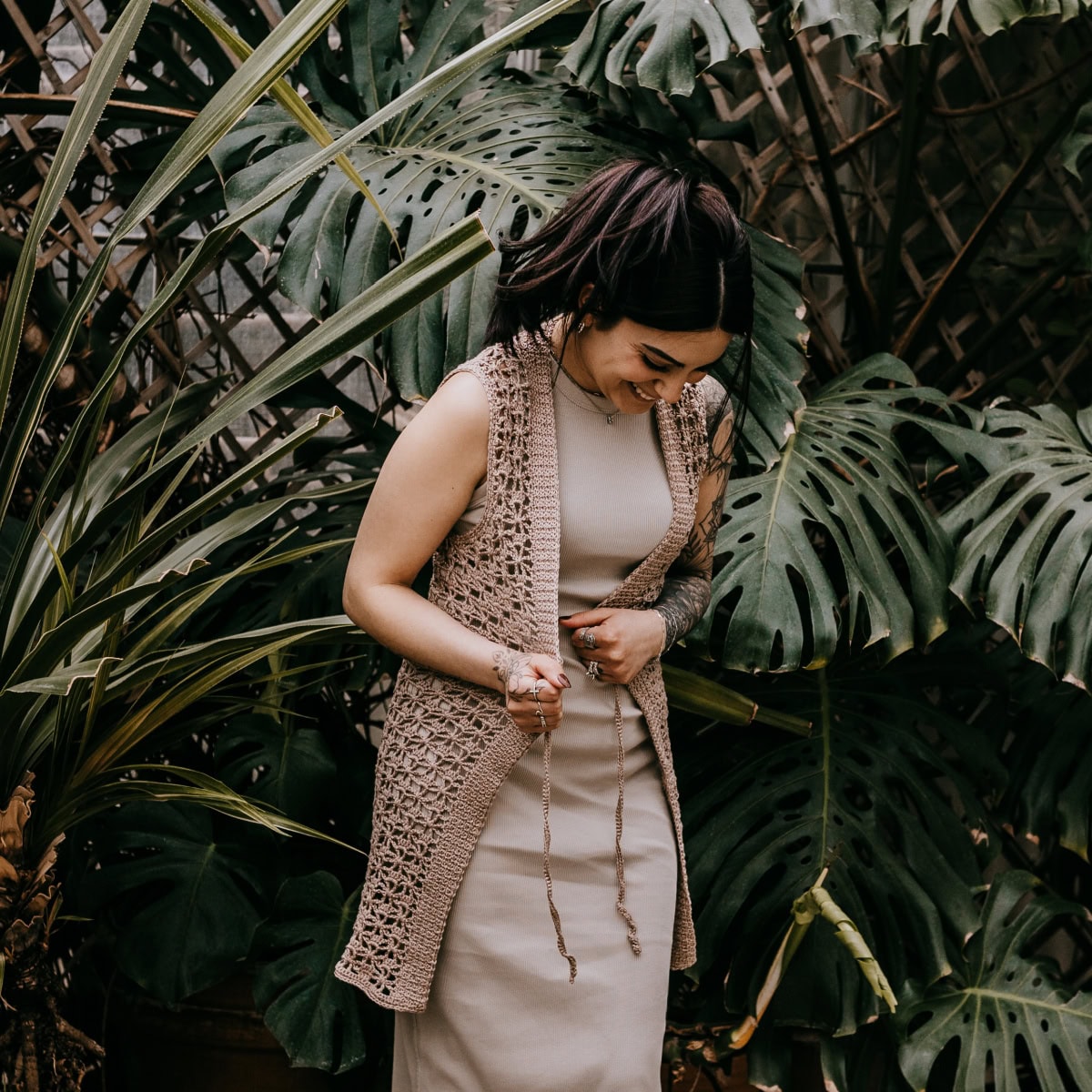 A young women wearing a long crochet vest over a knit dress standing in front of large greenery.