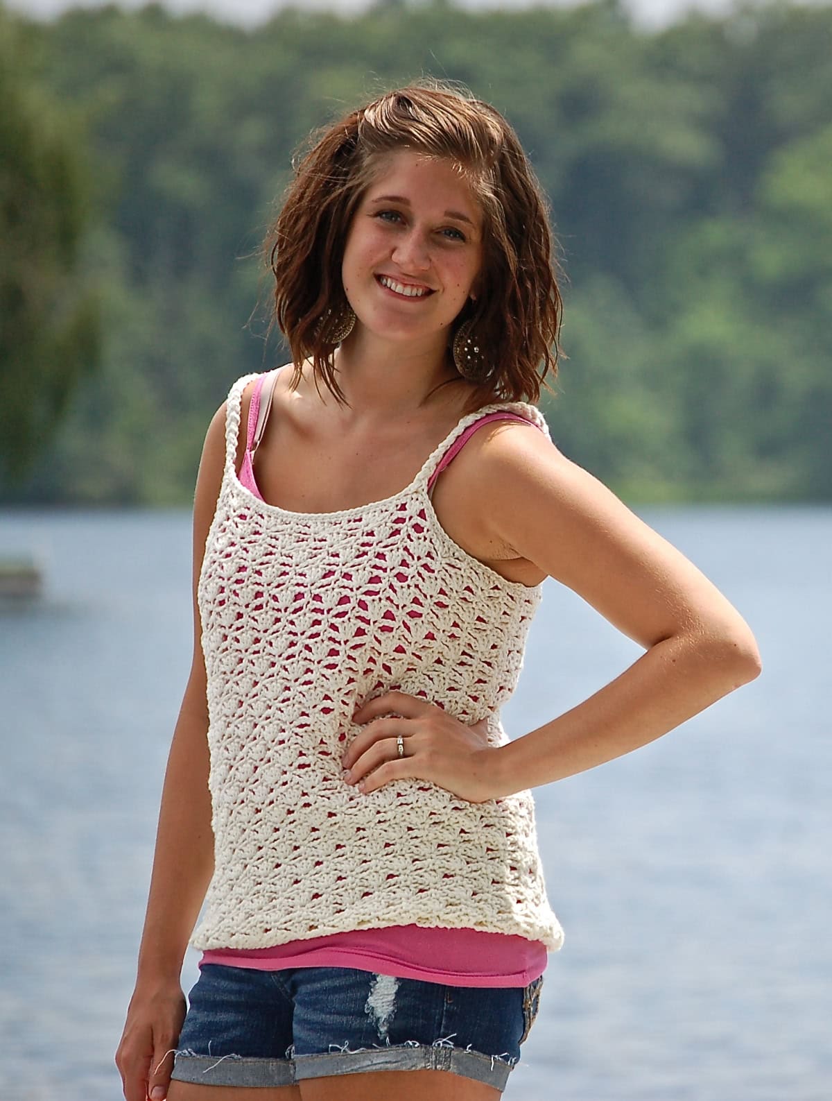 Woman posing with one hand on her waist wearing a white crochet tank top standing in front of a lake.