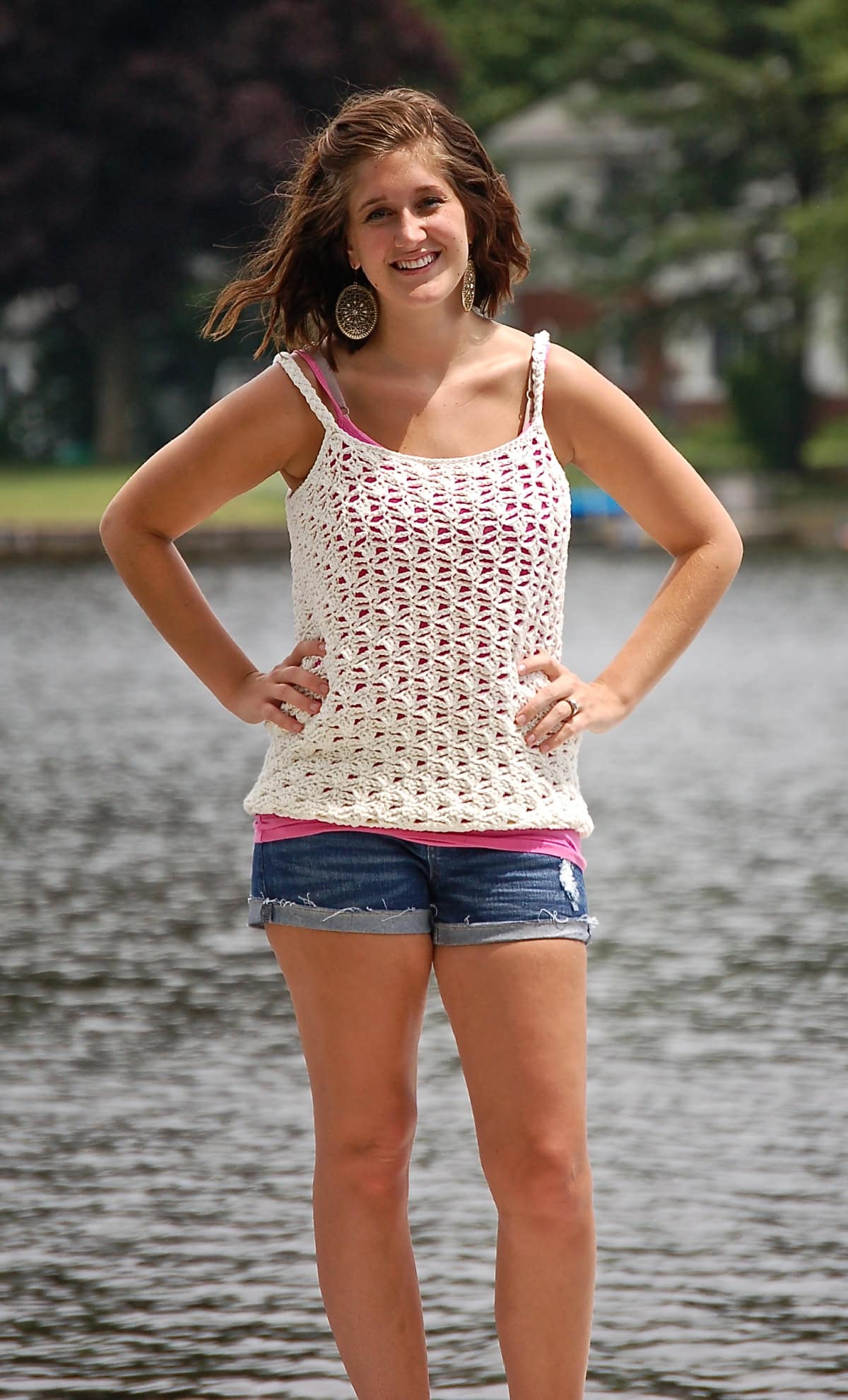 Woman wearing a white crochet tank top with her hands on her waist standing in front of a lake.