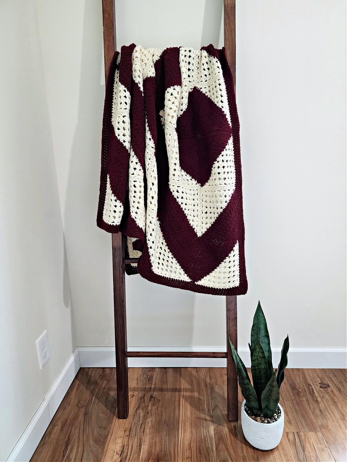 Granny squares crochet blanket hanging from a ladder standing on a wooden floor with a potted plant next to it.