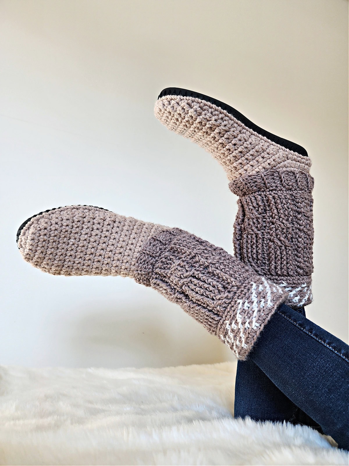 Brown crochet cable slipper boots in the air with a white sheet laid below.