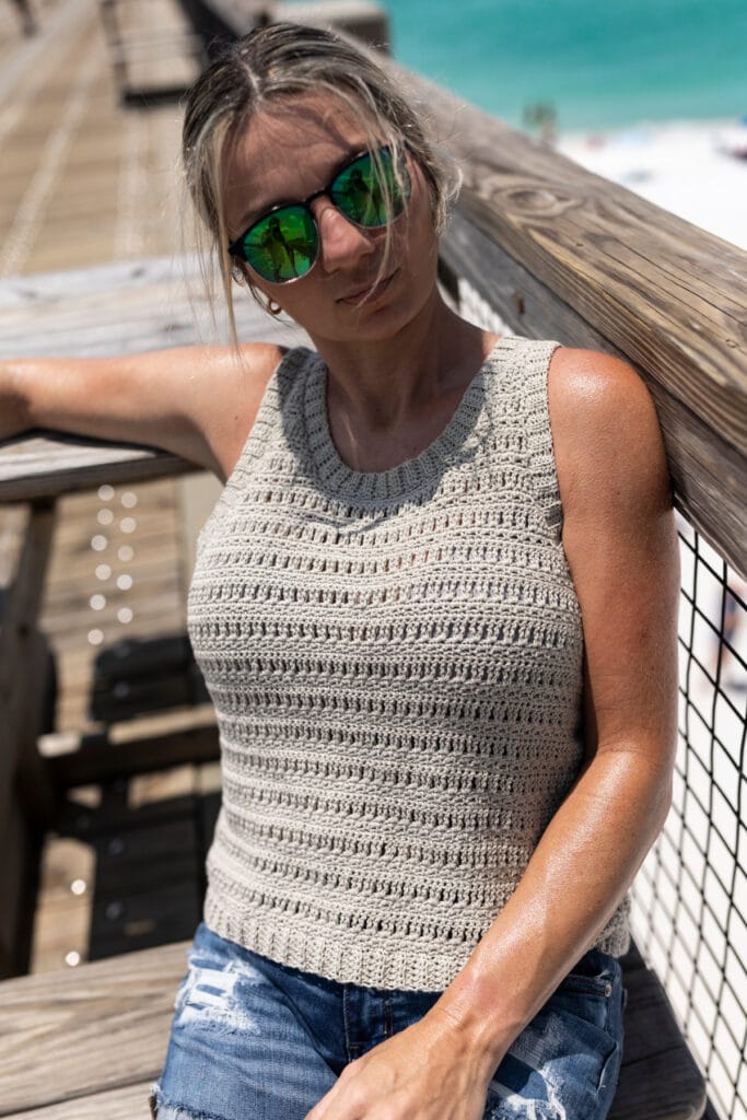 Woman sitting on a wooden bench at the beach wearing sunglasses and a crochet tank top.