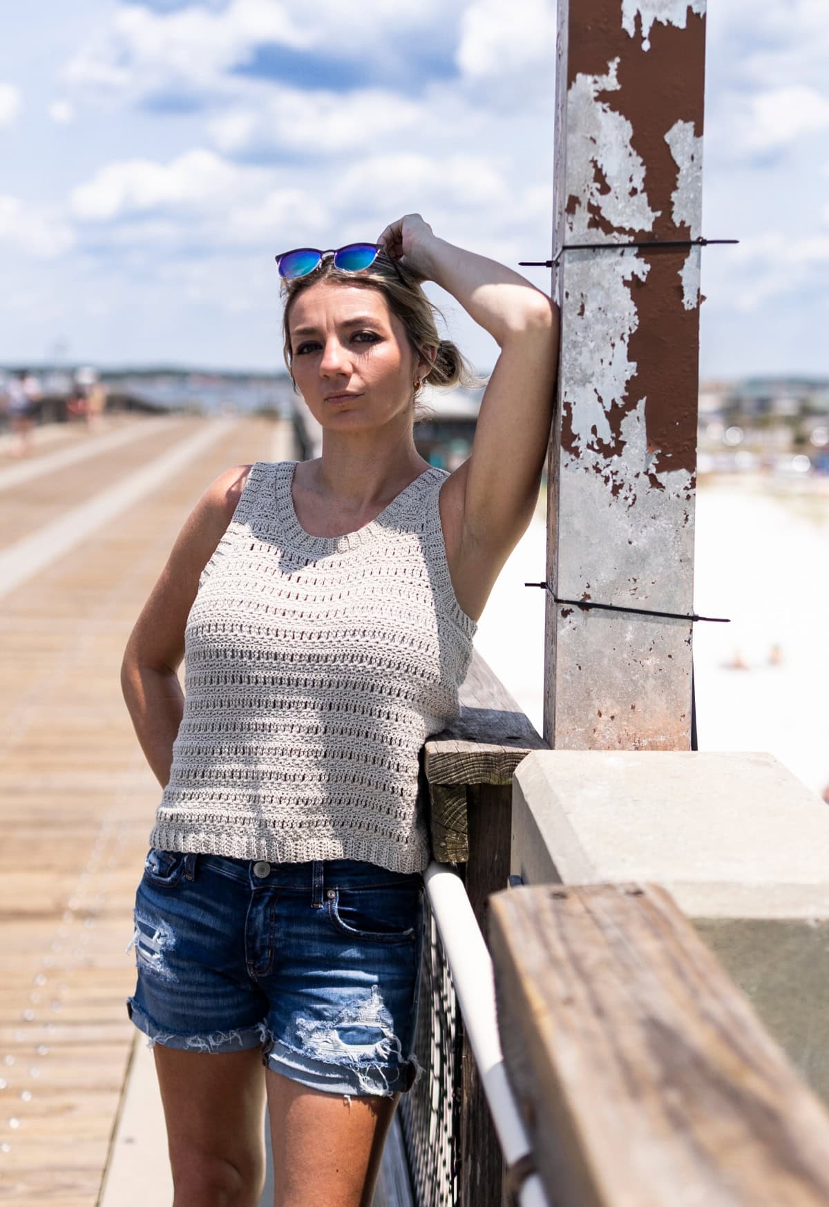 Woman leaning against a railing at the beach wearing sunglasses on top of her head and a crochet tank top with denim shorts.