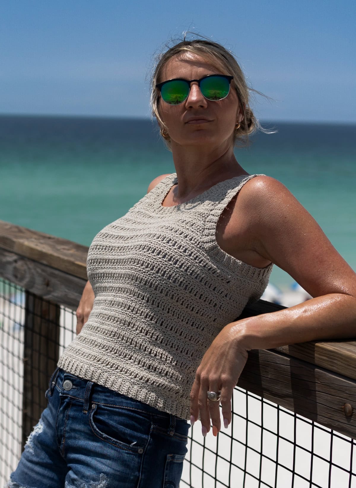 Woman leaning against a wooden railing at the beach wearing sunglasses and a crochet tank top.