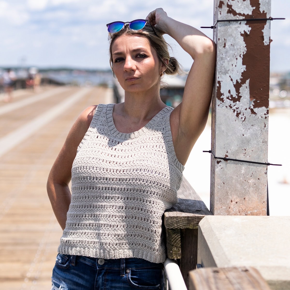 Woman leaning against a railing at the beach wearing sunglasses on top of her head and a crochet tank top with denim shorts.