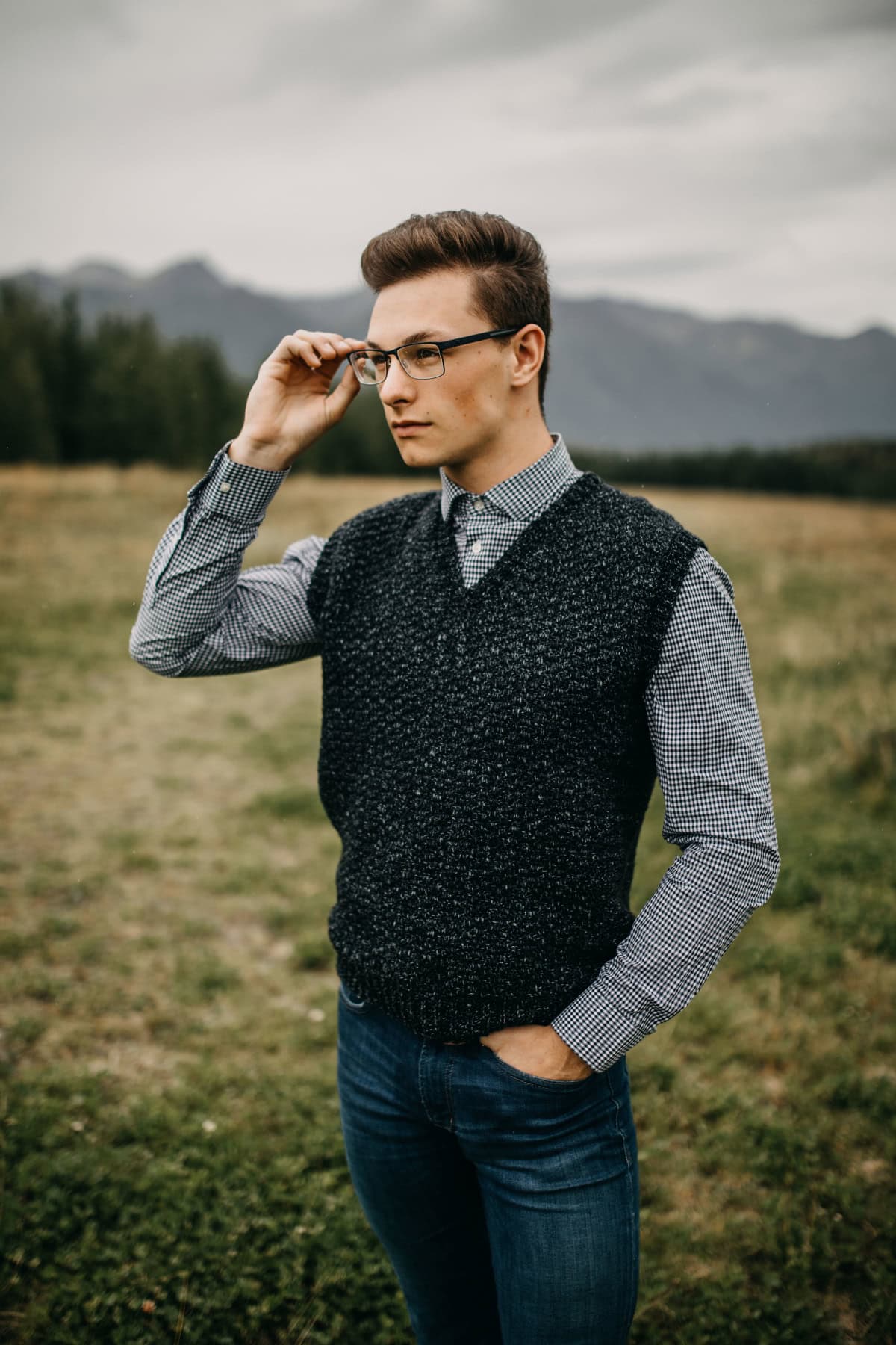 Man standing in a field wearing a dark crochet vest and glasses with one hand in his pocket and one hand on his glasses.