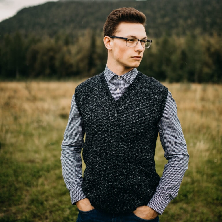 Man standing in a field wearing a dark crochet vest and glasses with his hands in his pockets.