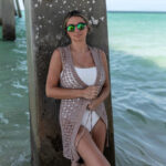 Woman wearing a crochet beach cover-up over a white swimsuit leaning against a pier in the ocean.