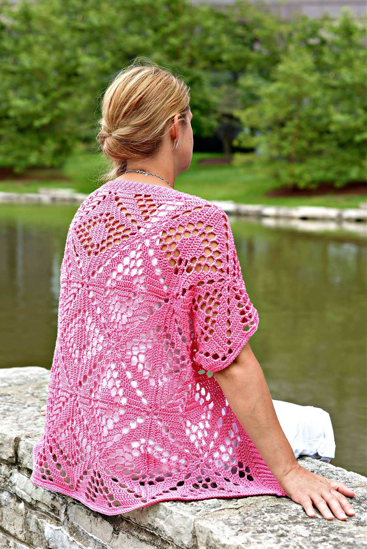 Woman sitting on a stone wall next to a lake wearing a pink lacy cardigan.