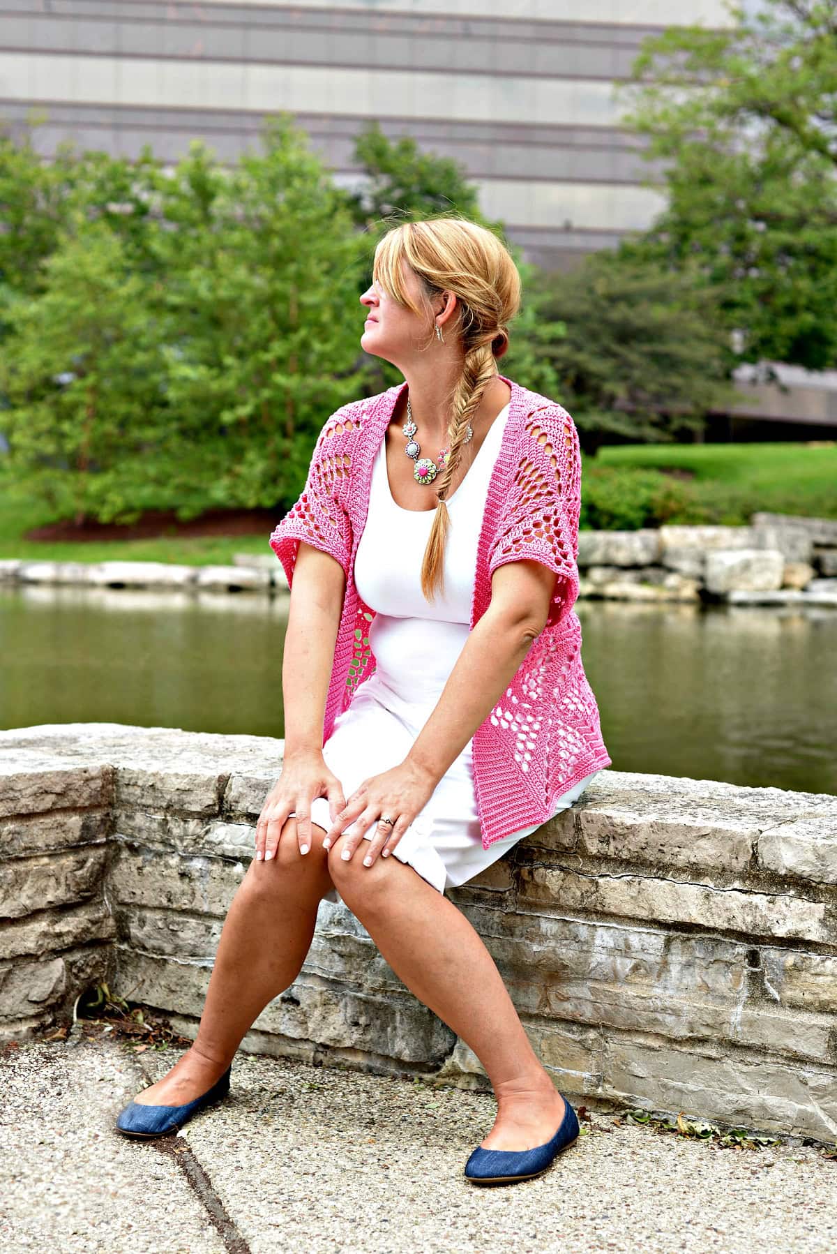 Woman sitting on a stone wall wearing a pink lacy cardigan and white shirt and shorts.