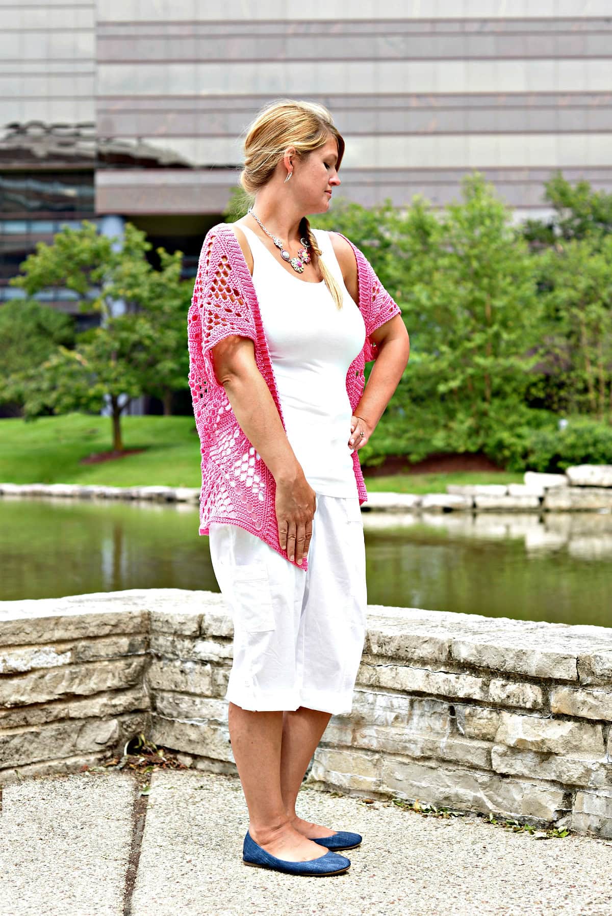 Woman standing in front of a stone wall wearing a pink lacy cardigan and white shirt and shorts.