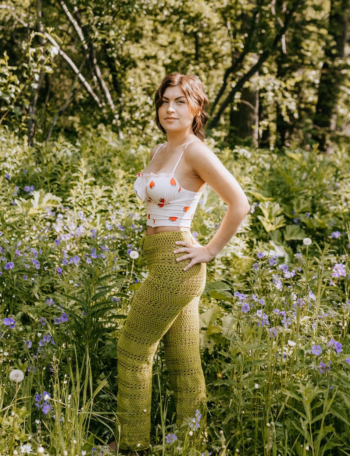 A woman wearing handmade green crochet pants and a white tank top standing in a field with one hand on her hip.
