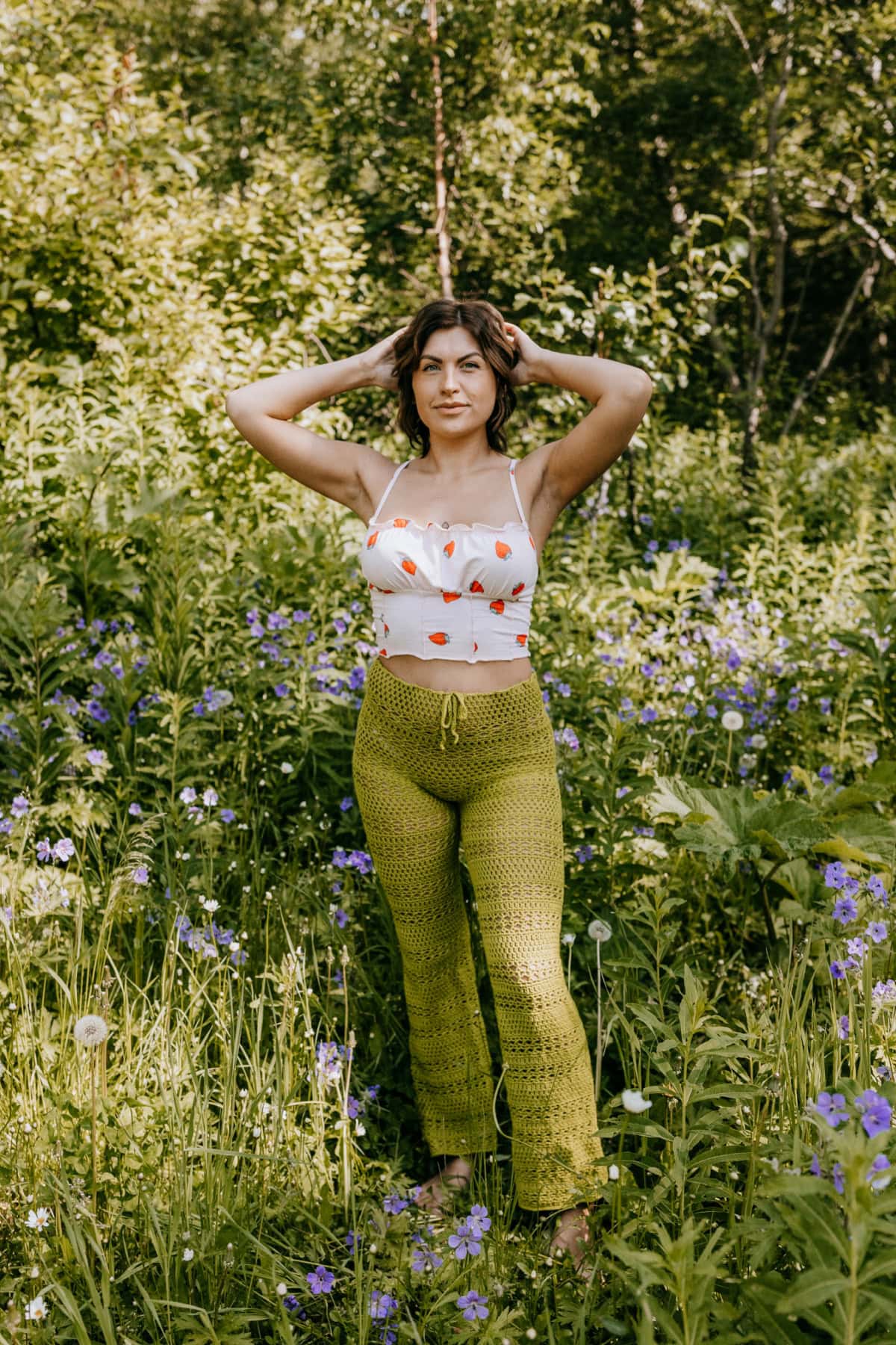 A woman wearing handmade green crochet pants and a white tank top standing in a field with her hands behind her head.