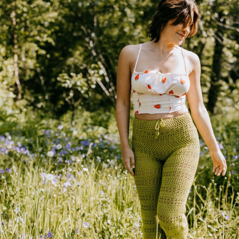 A woman wearing handmade green crochet pants and a white tank top standing in a field.