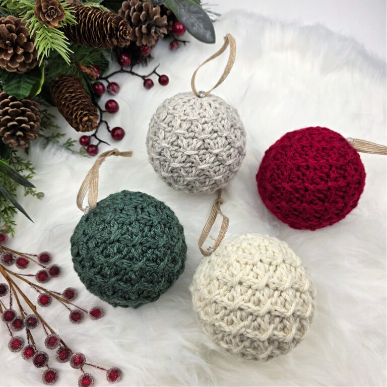 Knit-look crochet Christmas bauble laying on a faux fur rug with pinecones, berries, and greenery.