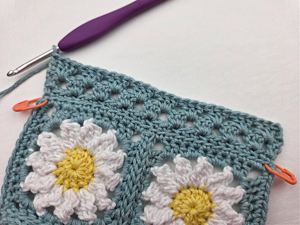 Working the border around two crochet daisy granny squares with a purple crochet hook.