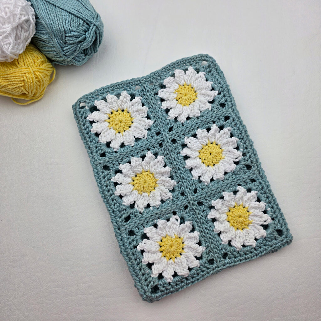 Six completed crochet daisy granny squares on a white surface with teal, yellow, and white yarn nearby.