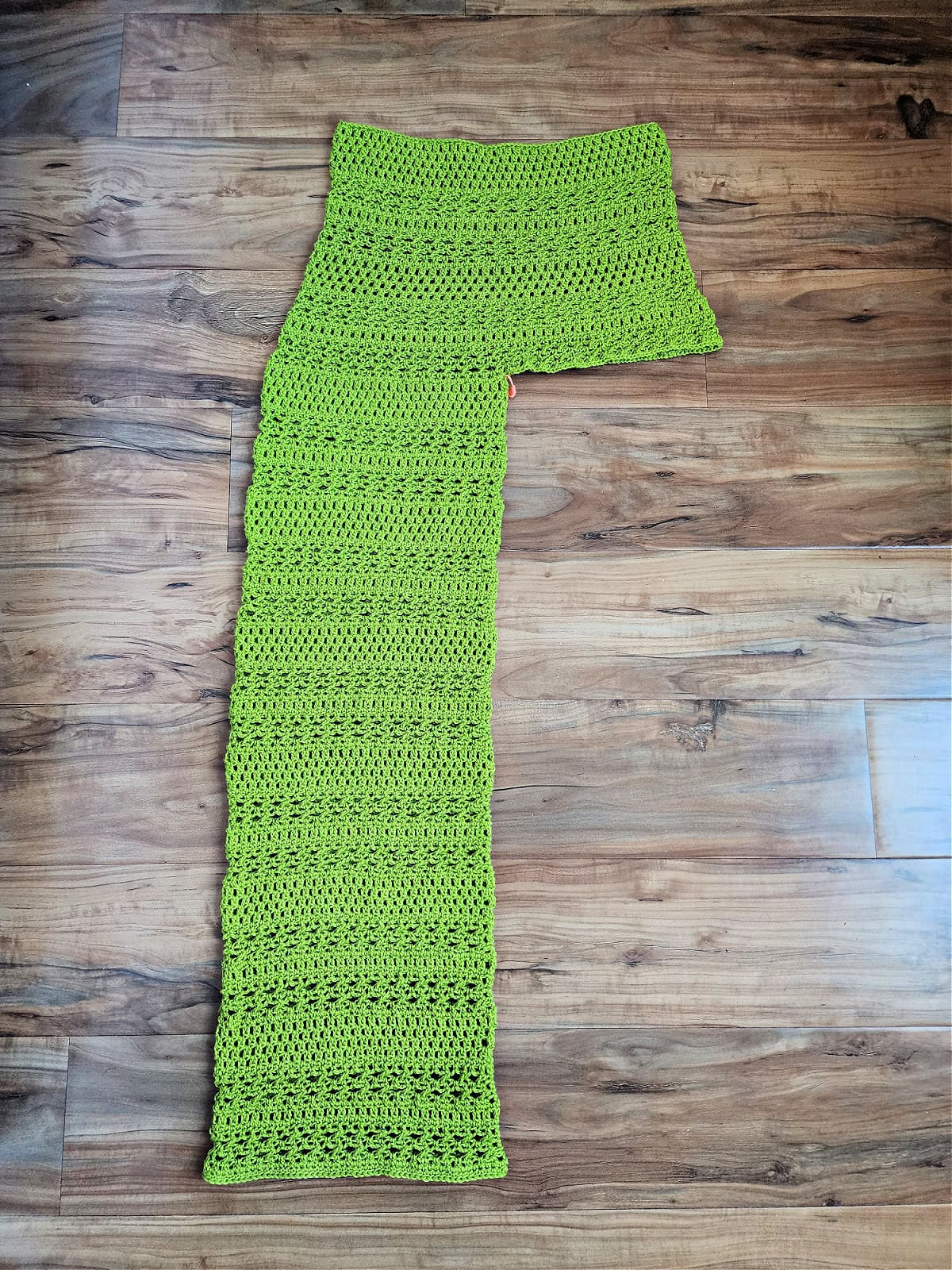 One completed leg of green crochet pants against a wooden floor.