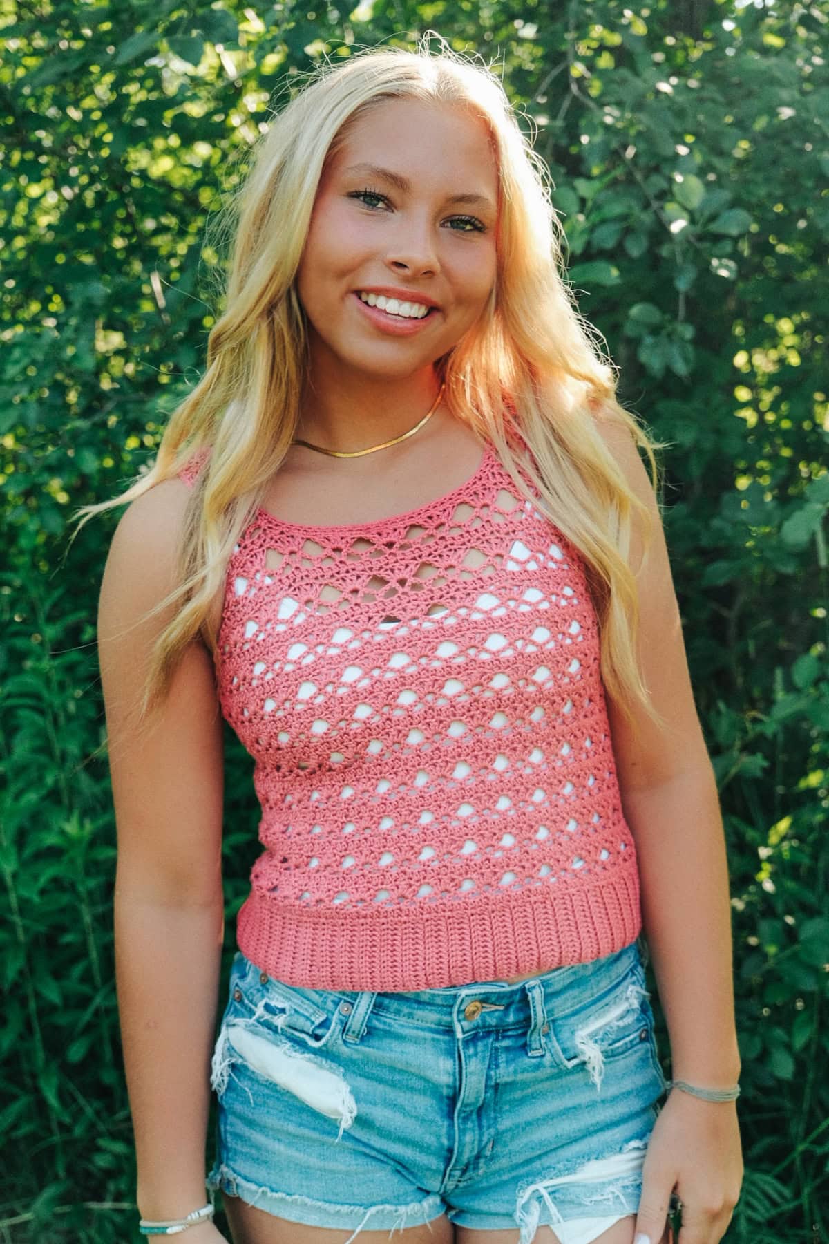 Woman wearing a red crochet tank top and blue denim shorts with trees in the background.