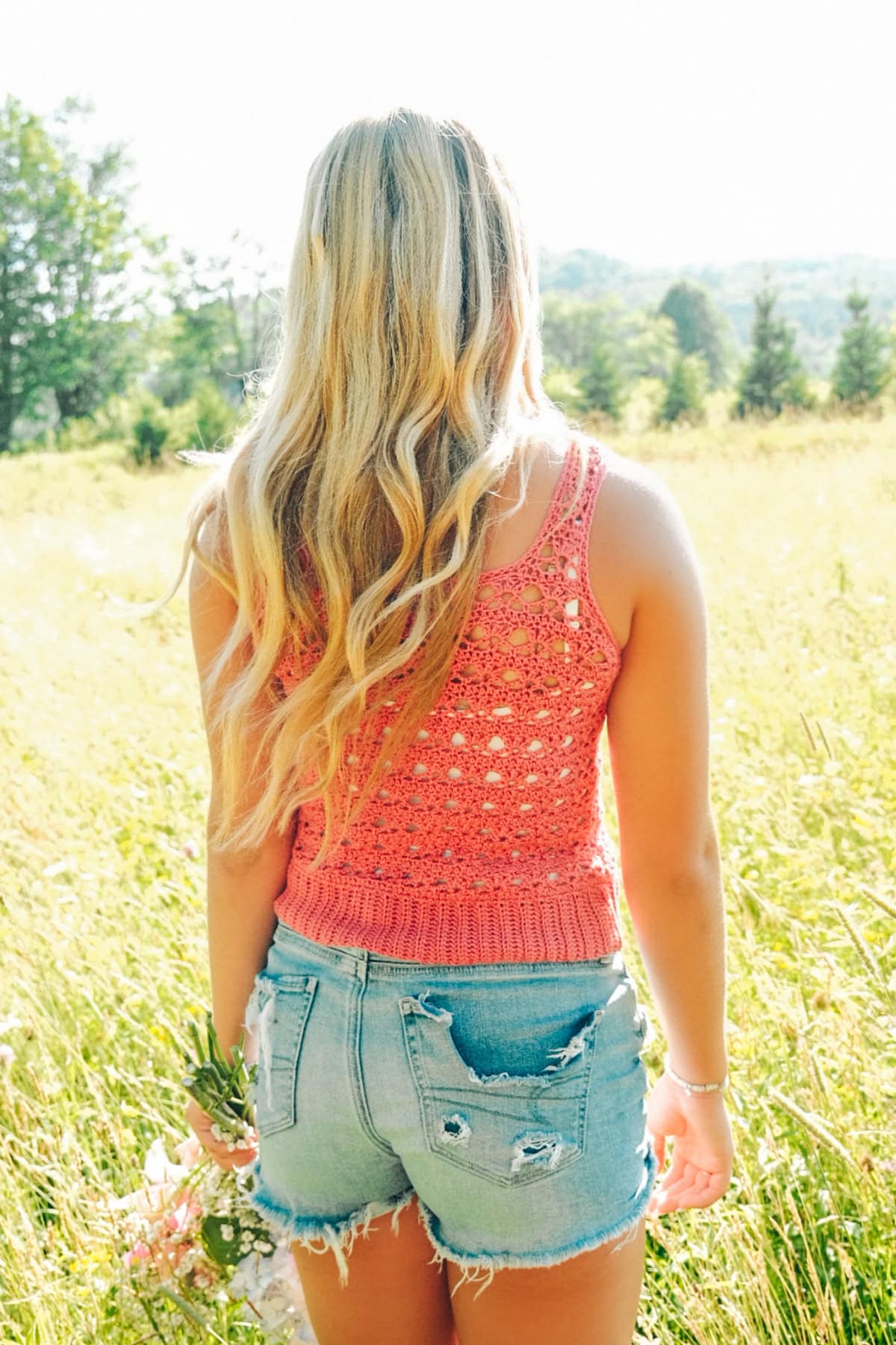 Woman wearing a red crochet tank top and blue denim shorts standing in a field with a bouquet in her hand and her back to the camera.