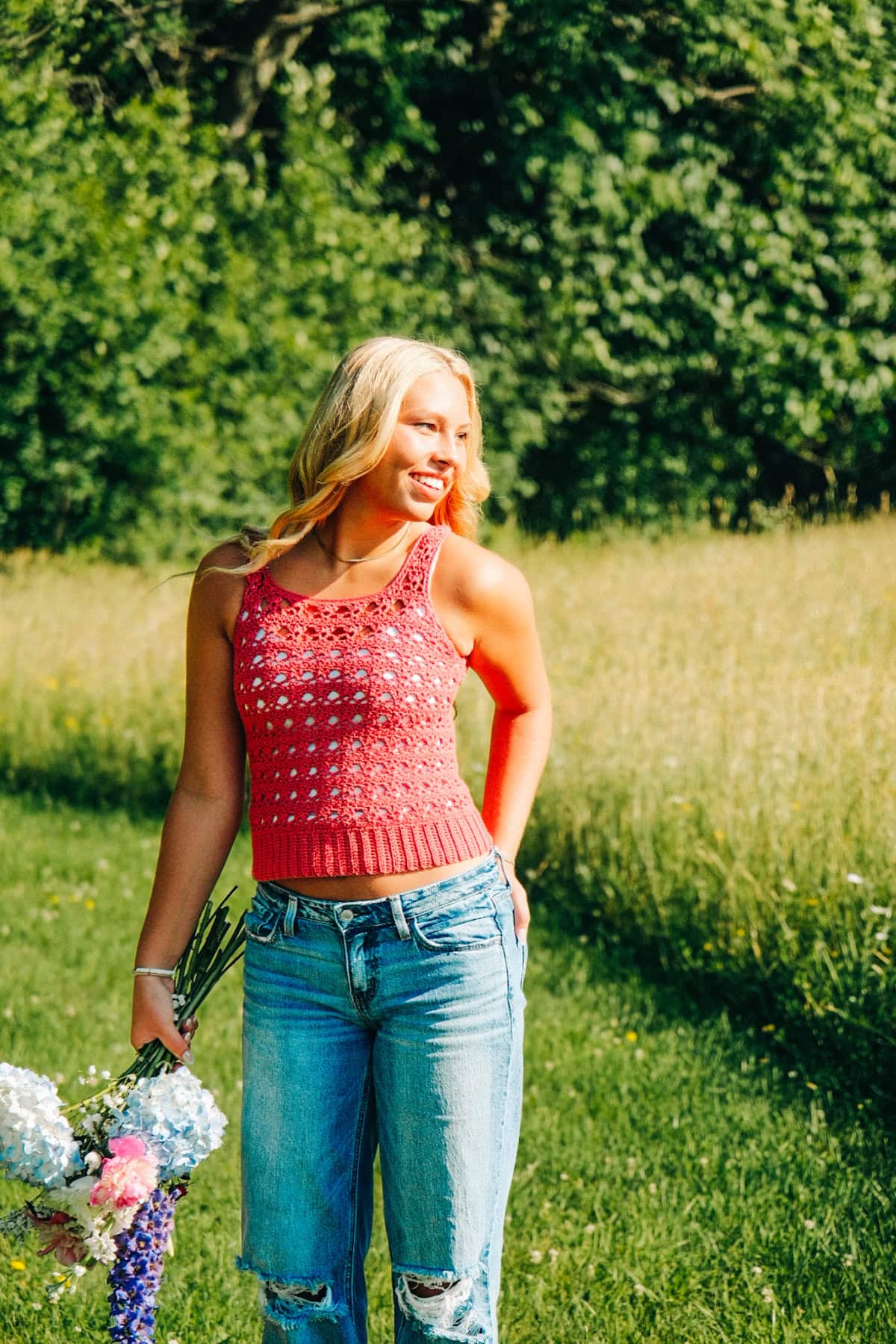 Woman wearing a red crochet tank top and blue jeans standing in a field with a bouquet in her hand.