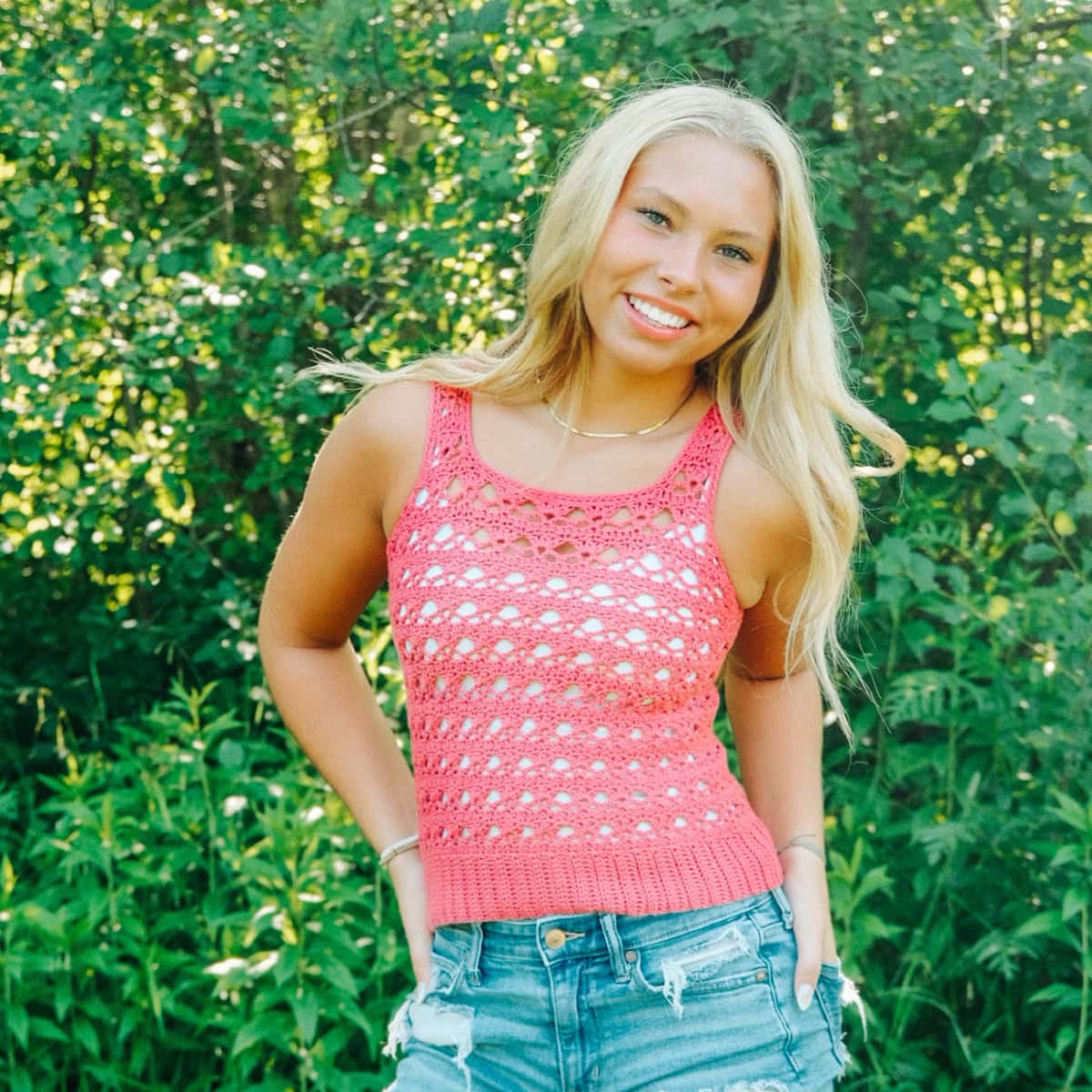 Woman wearing a red crochet tank top and blue denim shorts with trees in the background and her hands on her hips.