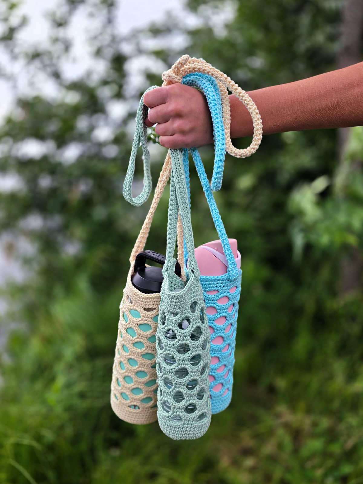 A hand holding off-white, green, and blue colored crochet water bottle holders outdoors.