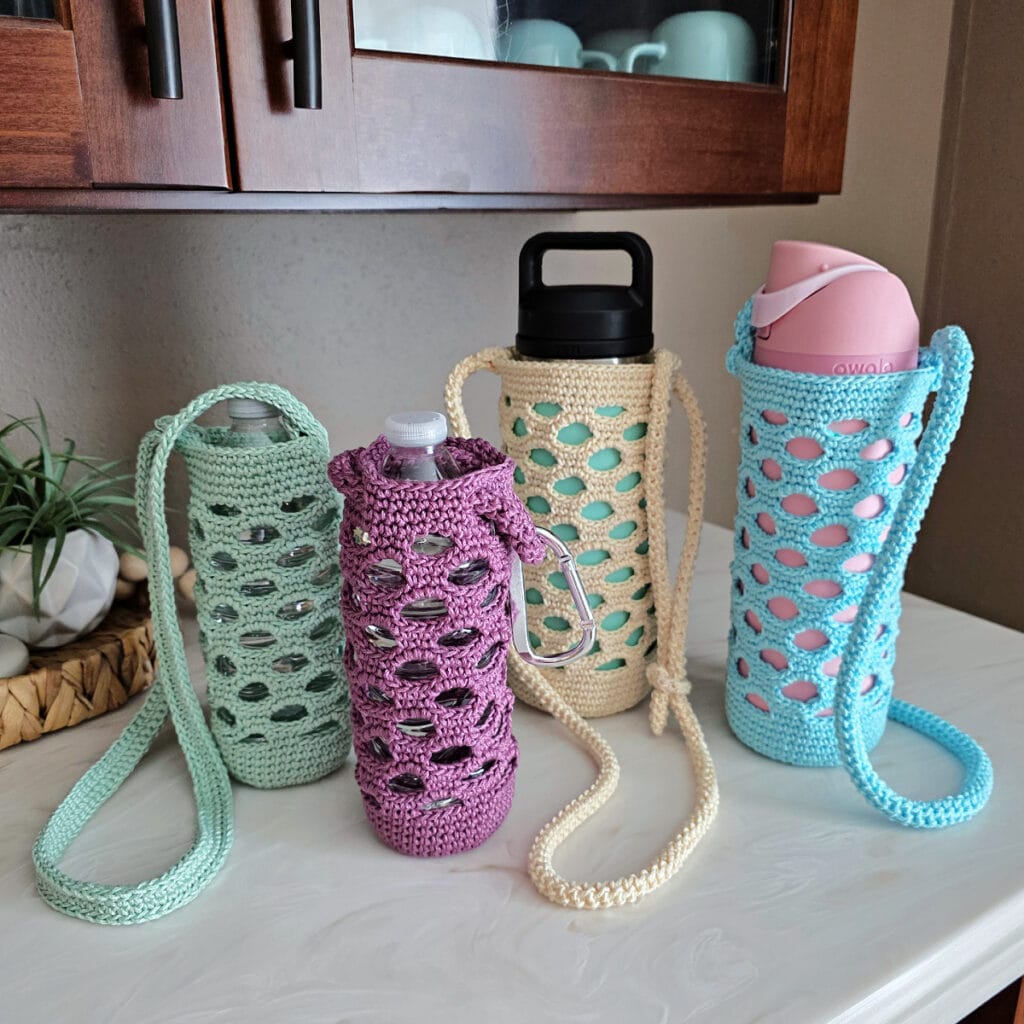 Green, purple, off-white, and blue colored crochet water bottle holders on a white counter.
