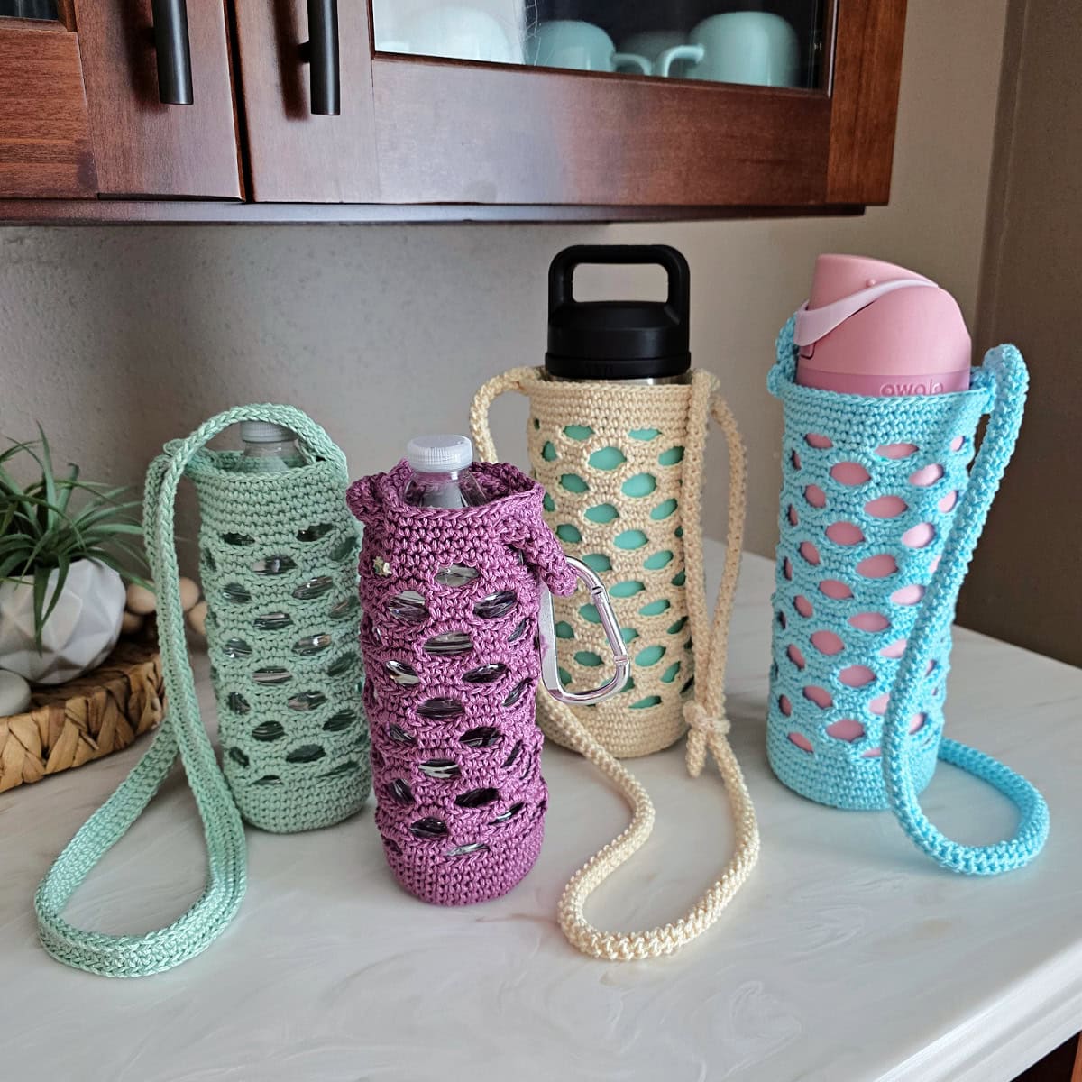 Green, purple, off-white, and blue colored crochet water bottle holders on a white counter.