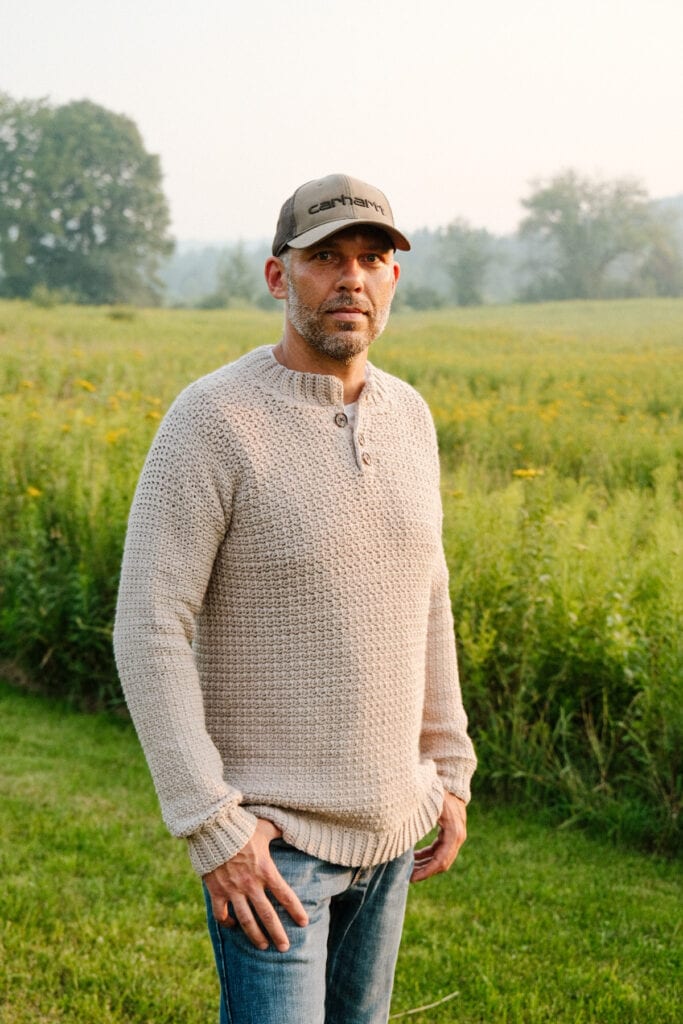 A man standing in a field wearing a beige crochet sweater and hat.