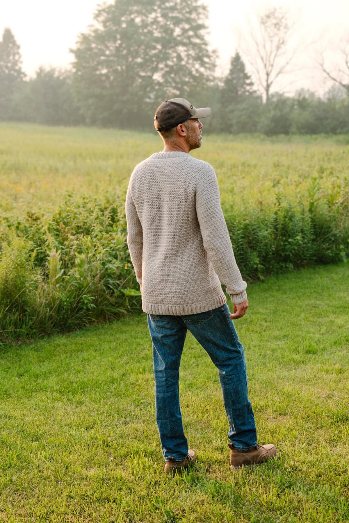 A man standing in a field with his back to the camera wearing a beige crochet sweater and hat.