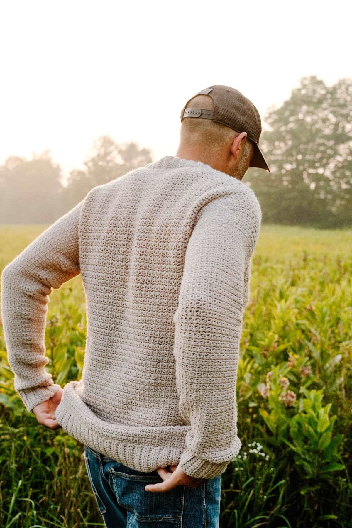 A man standing in a field wearing a beige crochet sweater and hat with his back to the camera.