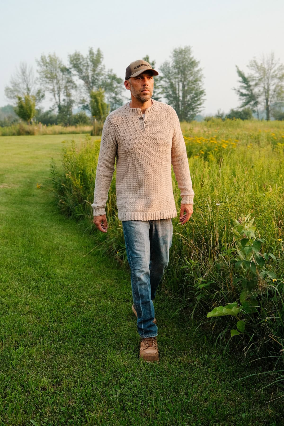 A man walking through a field wearing a beige crochet men's sweater, blue jeans and hat.
