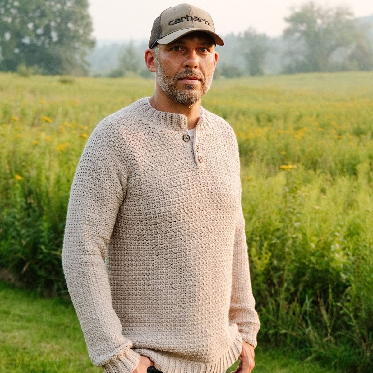 A man standing in a field wearing a beige crochet sweater and hat.