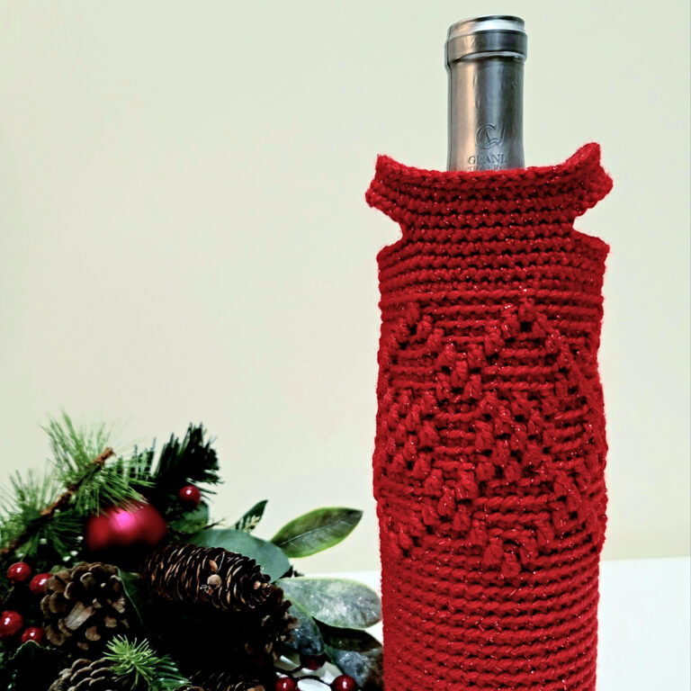 An argyle crochet wine tote in red sparkly yarn sitting next to holiday greenery.