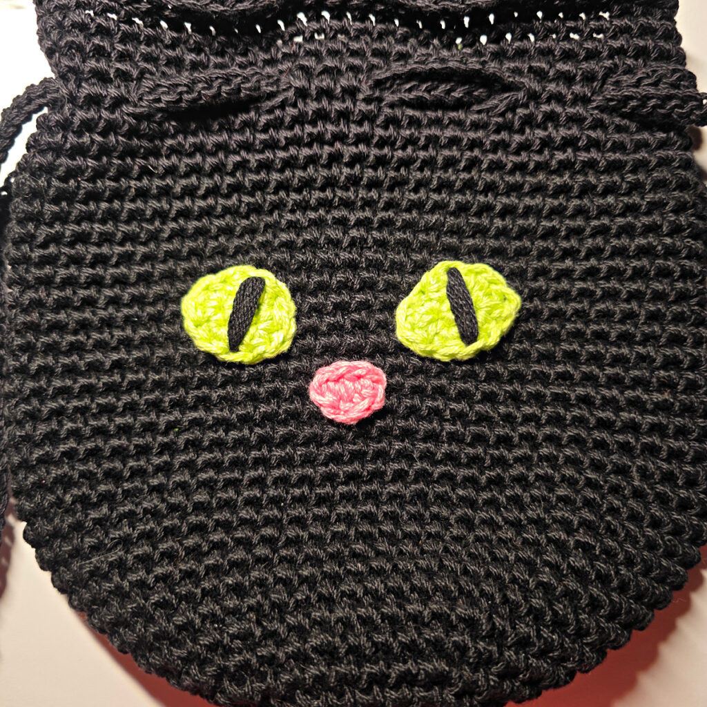 A black cat crochet treat bag with a pink nose and green eyes.