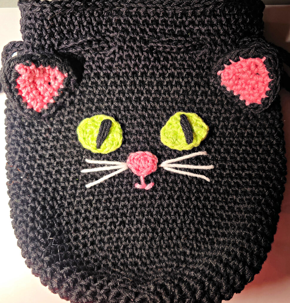 A black cat crochet treat bag with a pink nose, ears and green eyes.