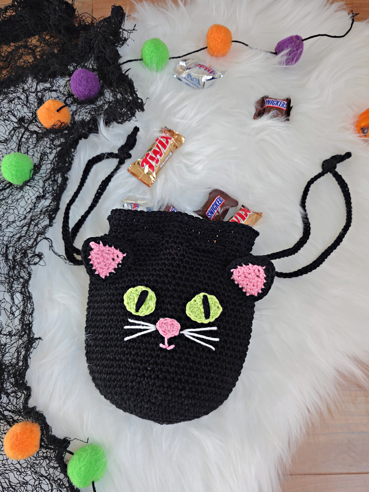 A black cat crochet treat bag full of candy against a white faux fur and web background.