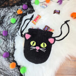 A black cat crochet treat bag full of candy against a white faux fur and web background.