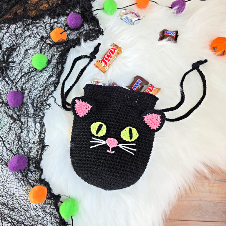 A black cat crochet treat bag full of candy against a white faux fur and web background.