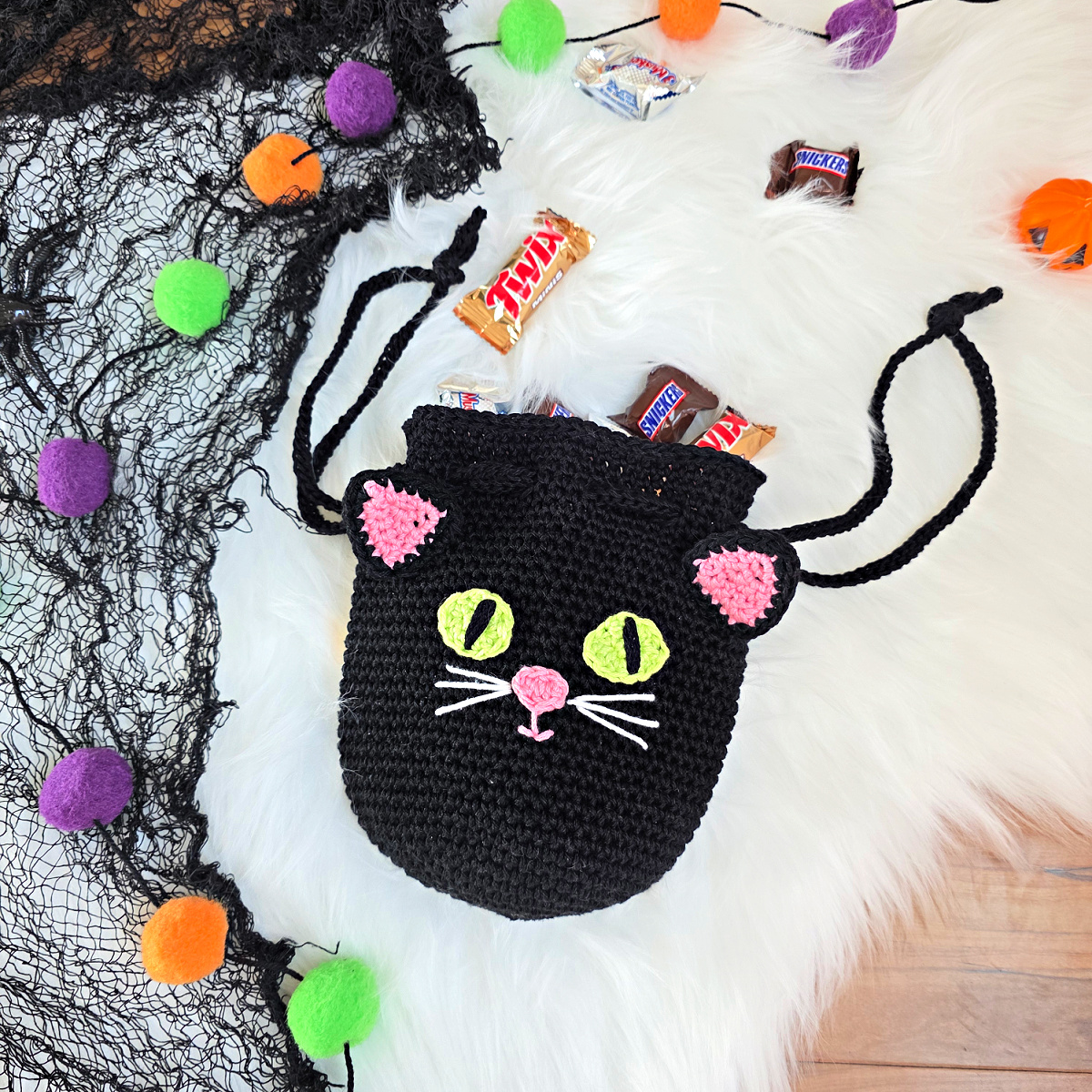 A black cat crochet treat bag full of candy against a white faux fur and web background.