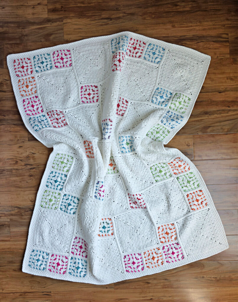 White crochet blanket with colorful squares spread out over the wooden floor.