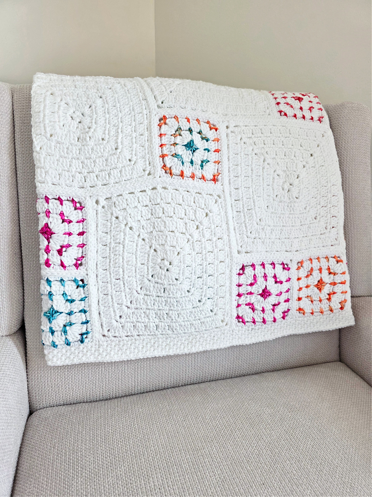 White crochet blanket with colorful squares folded over a sofa.