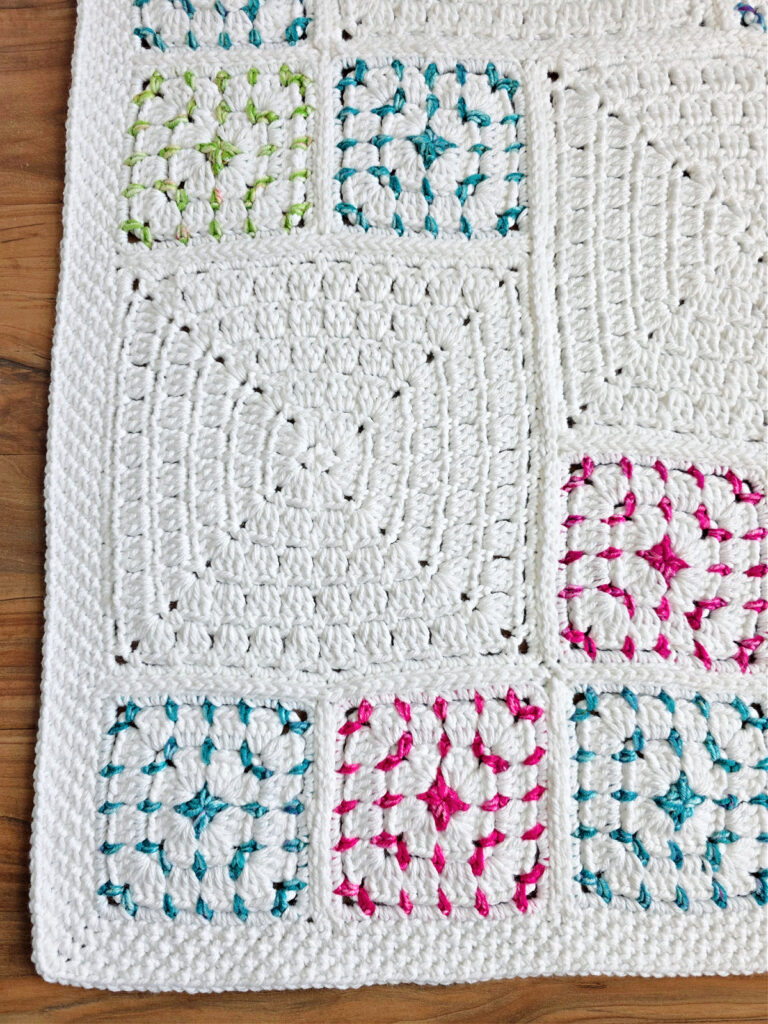 Corner of a white crochet blanket with colorful squares spread out over a wooden floor.
