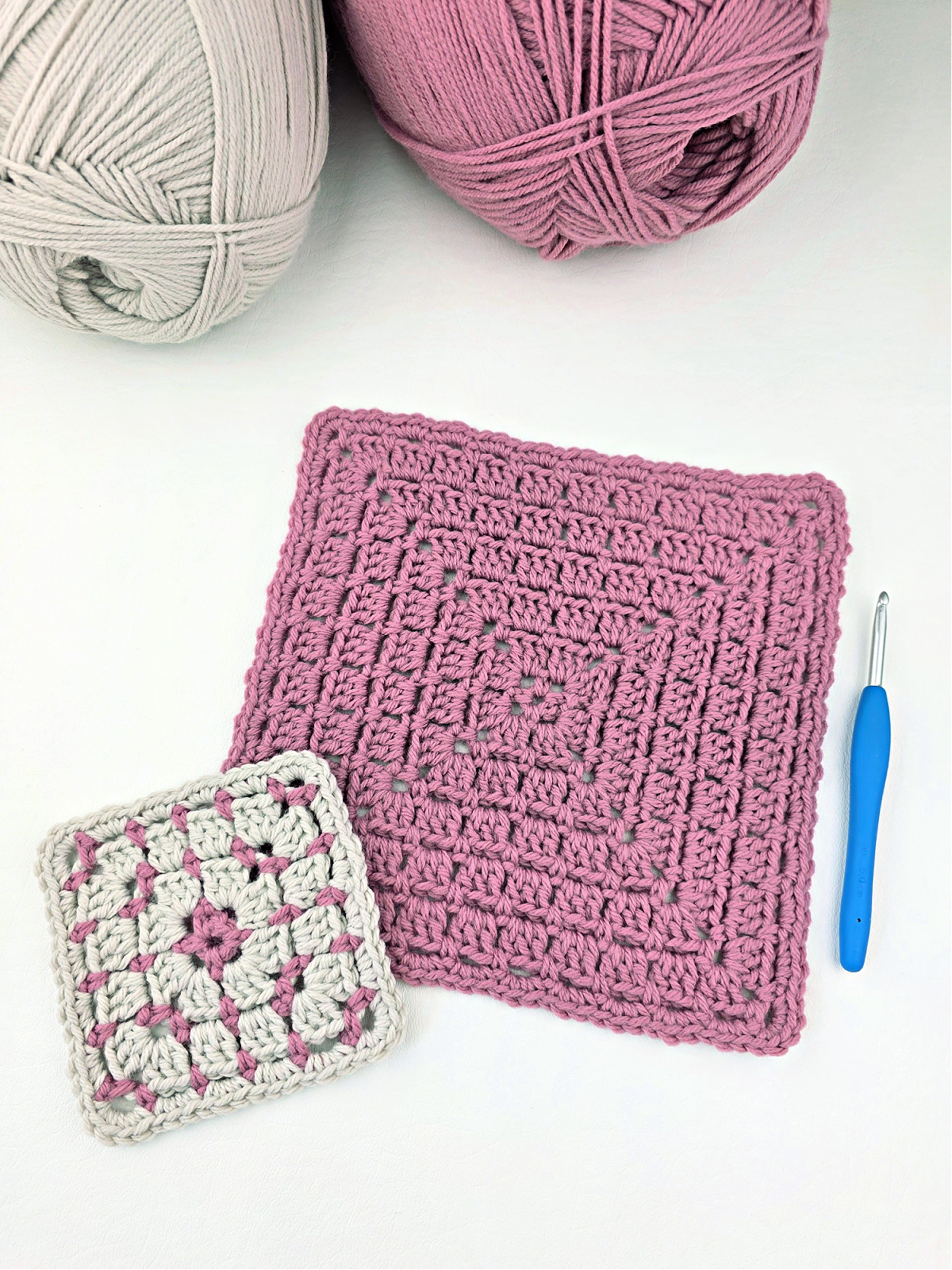 One large pink block stitch granny square and one small white and pink granny square with two balls of yarn in the corner and a blue crochet hook.