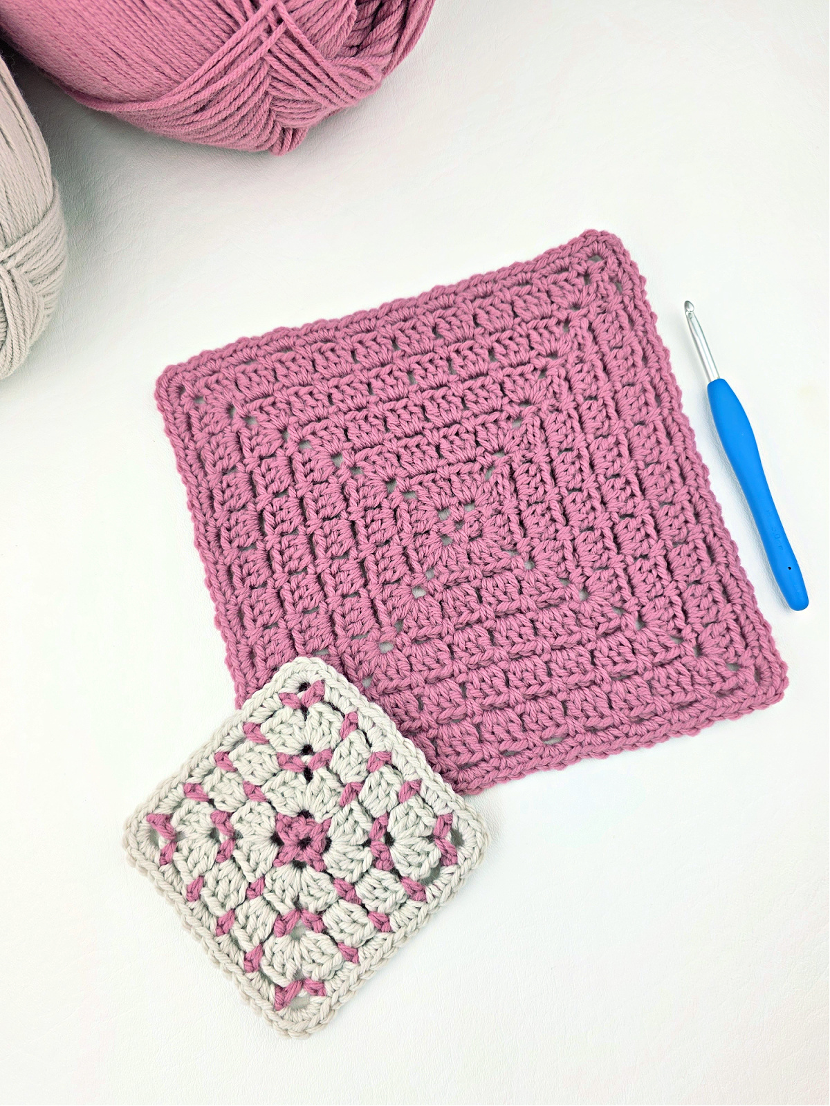 One large pink block stitch granny square and one small white and pink granny square with two balls of yarn in the corner and a blue crochet hook.