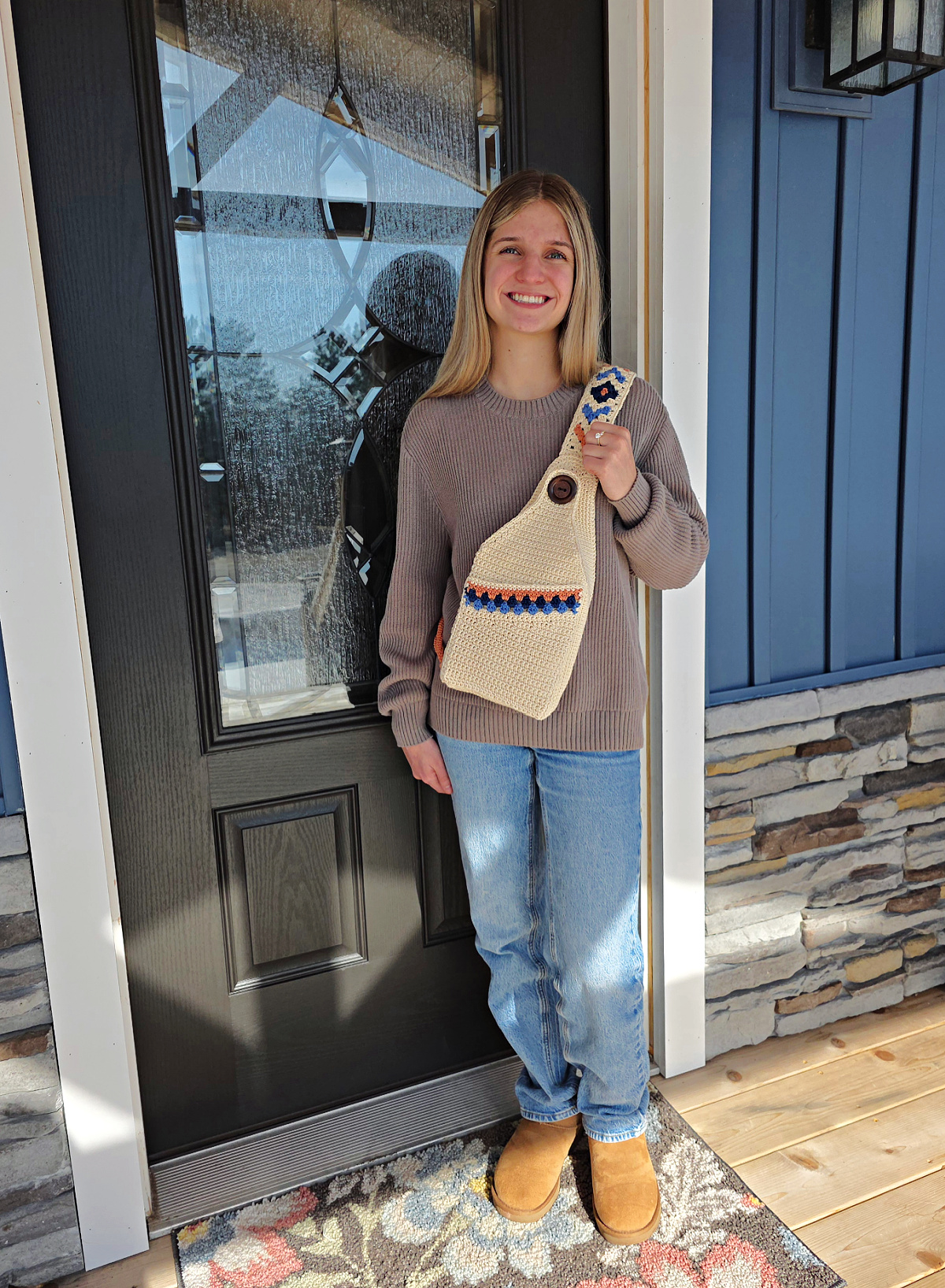 Girl in a brown pullover with a boho crochet sling bag over her shoulder standing next to a door.