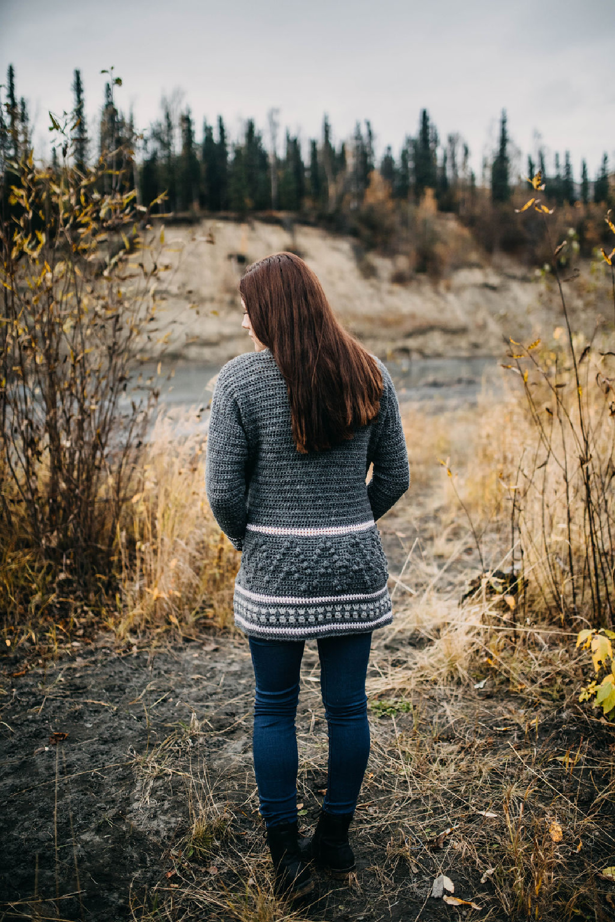 Woman wearing a grey crochet cardigan with black jeans standing in a field with her back to the camera.