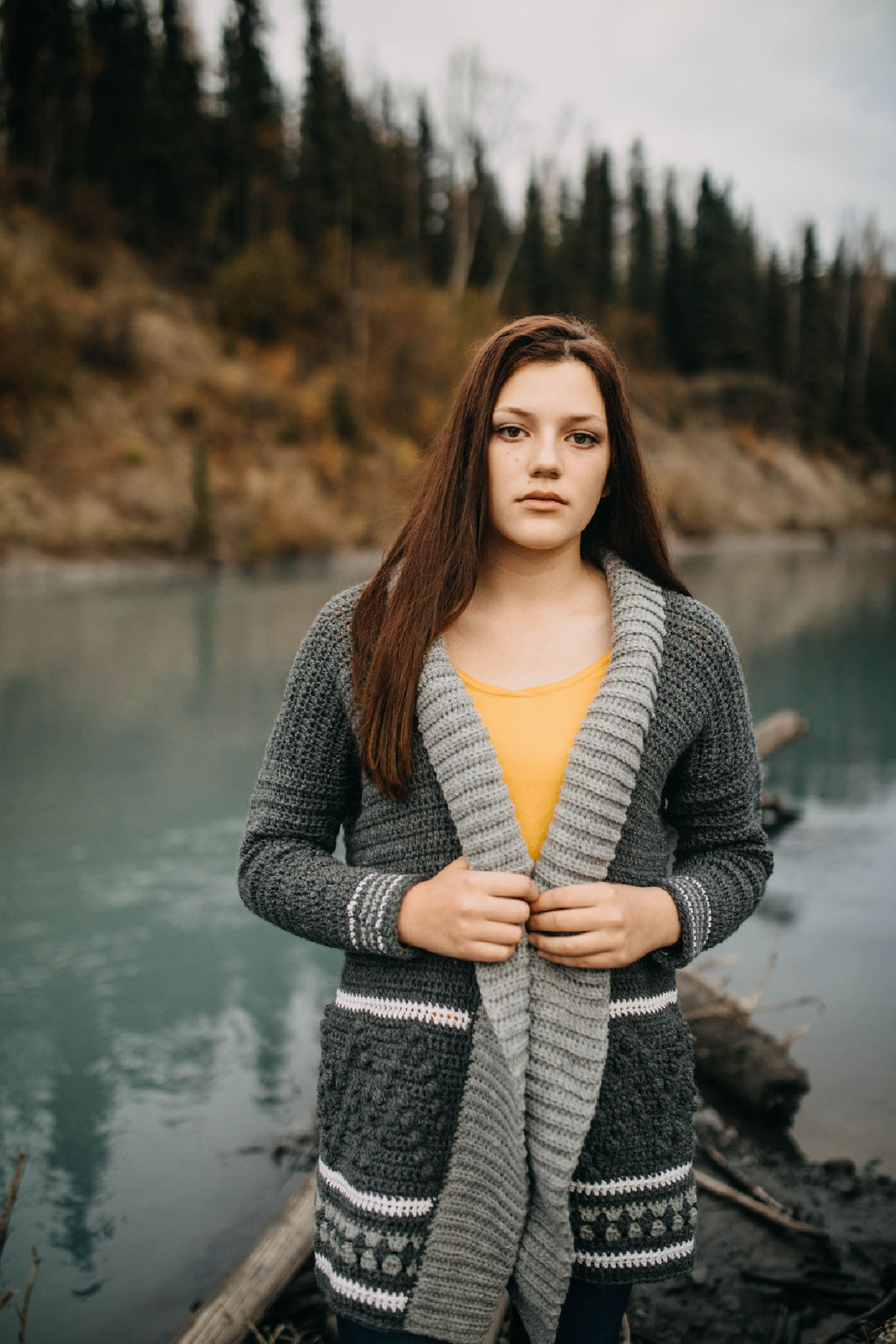 Woman wearing a grey crochet cardigan with a yellow top standing in front of a water body.